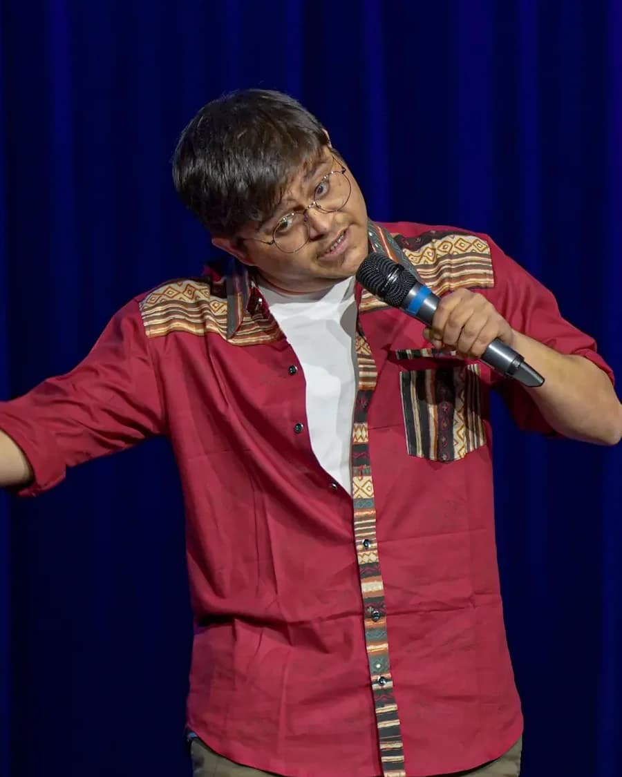 Om Bhatt performing with a microphone in a red patterned shirt against blue stage curtains